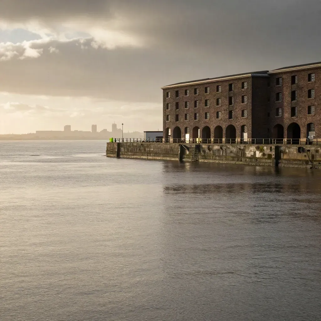 Liverpool dock edges, pale estuary water, warehouse arches and skyline in hazy cloud-break light.